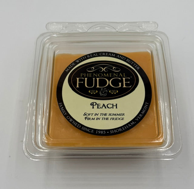 A square tub of peach-flavored fudge in a clear plastic clamshell container with a circular black-and-cream label reading Phenomenal Fudge peach.
