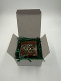 A square piece of chocolate fudge wrapped in clear cellophane with a gold and black label reading 'Phenomenal Fudge,' displayed inside an open white gift box filled with green crinkle paper on a white background.