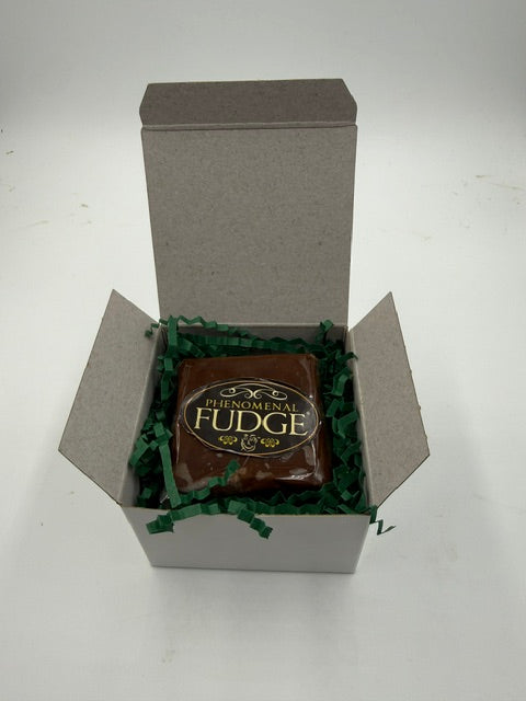A square piece of chocolate fudge wrapped in clear cellophane with a gold and black label reading 'Phenomenal Fudge,' displayed inside an open white gift box filled with green crinkle paper on a white background.